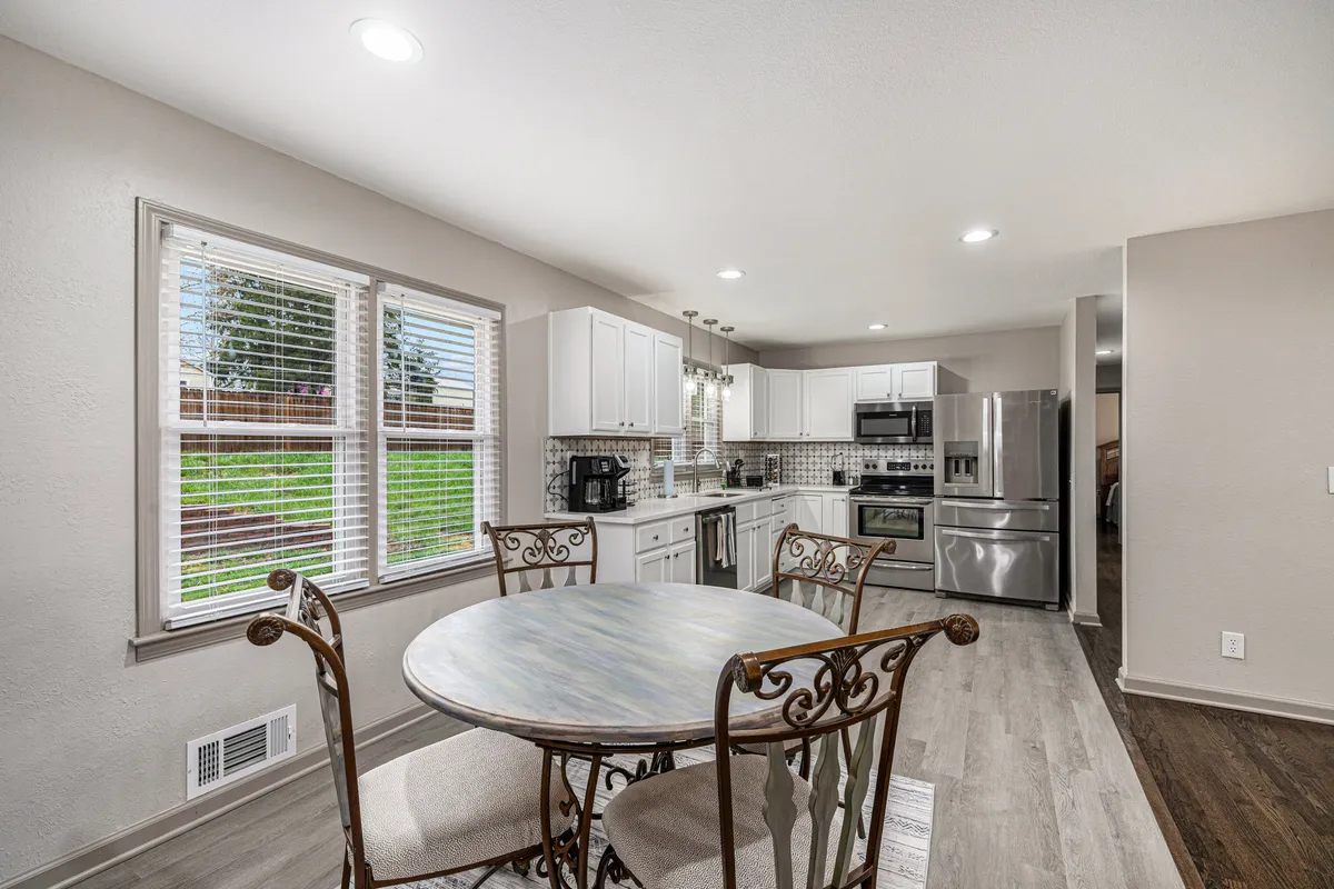 This image presents a warm, welcoming view of a round dining table adjacent to a kitchen island, with large windows allowing natural light to fill the space. The scene is set for family meals or casual gatherings, and the layout clearly encourages both cooking and socializing. The visible openness between kitchen and dining area echoes the open-concept layout highlighted in the section.