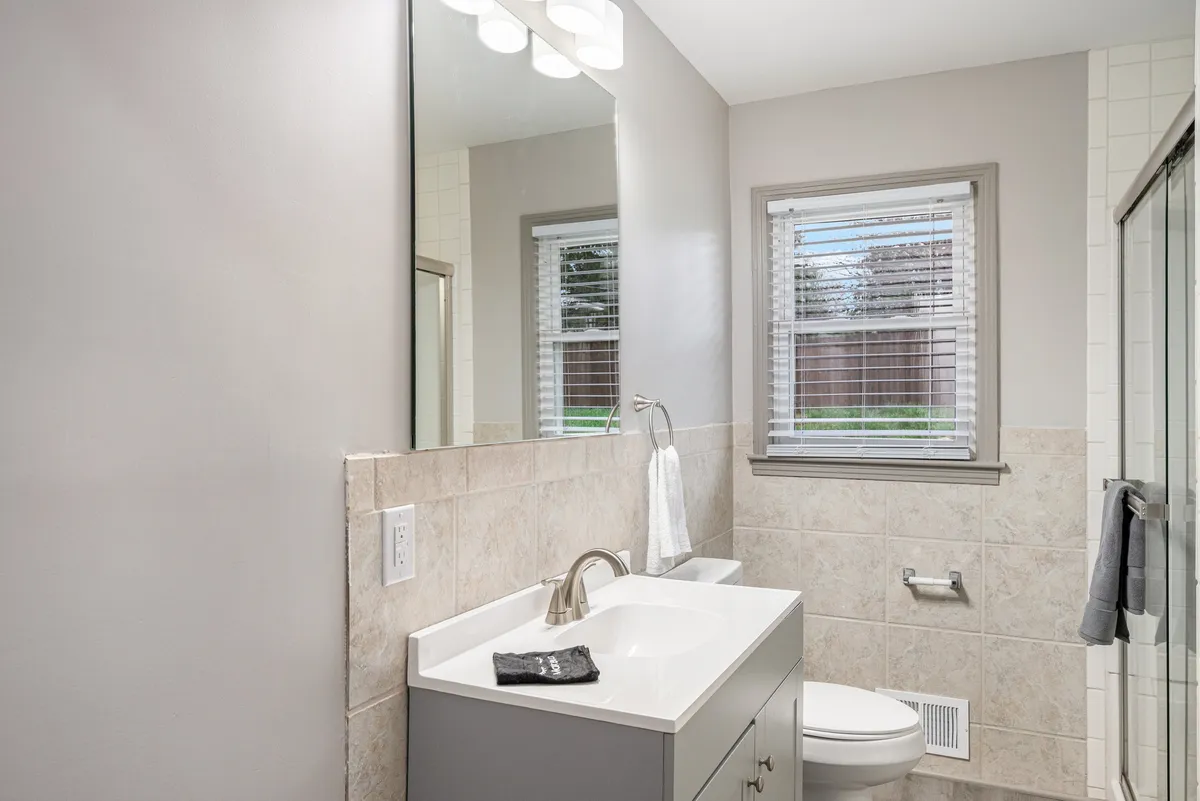 A well-lit bathroom with a modern vanity, a large mirror, and a window allowing in fresh morning sunlight. The setting feels fresh and inviting, ideal for personal grooming or a refreshing shower after a long day.