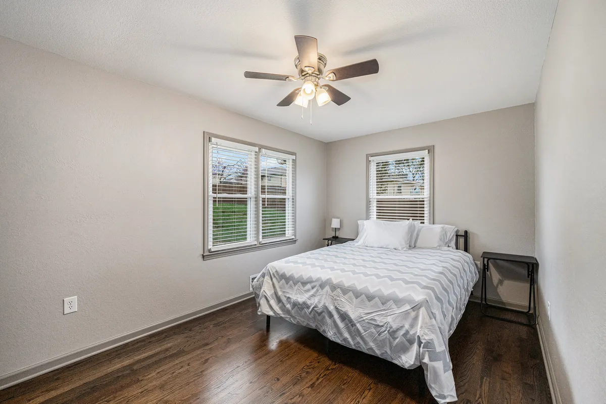 A serene bedroom scene featuring a bed with a modern chevron-patterned duvet, soft natural light streaming in through windows with blinds, and a ceiling fan overhead. The overall mood is tranquil, suggesting a restful atmosphere ideal for unwinding after a busy day.