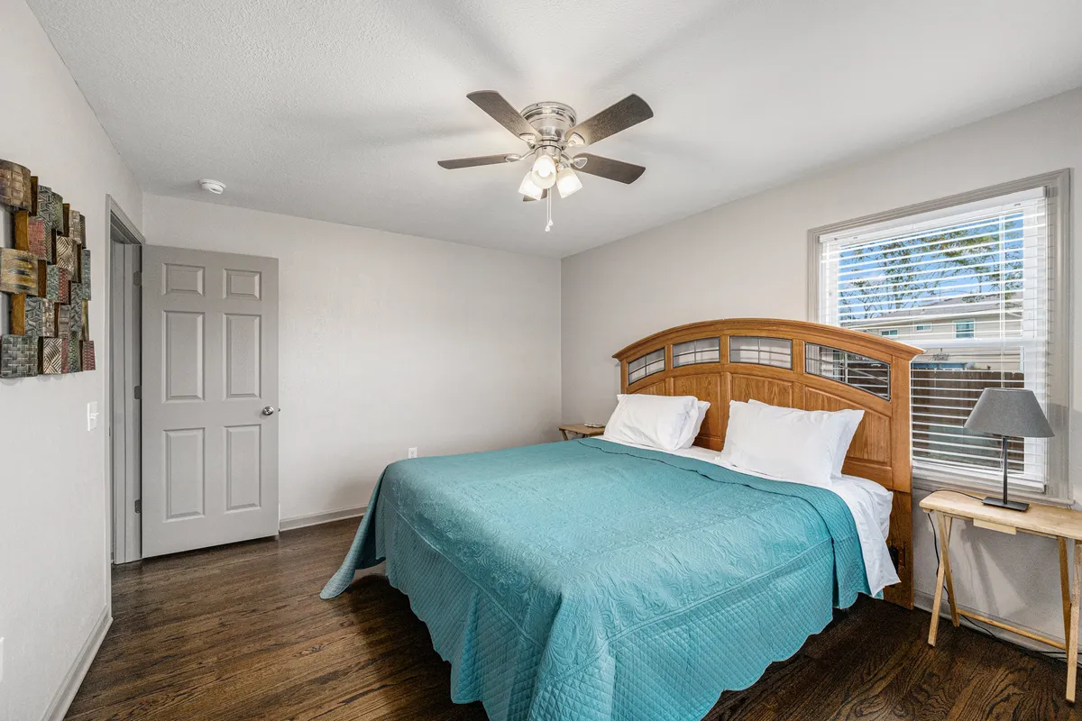 A serene bedroom featuring a bed with a plush teal quilt, wooden headboard, cozy side table with lamp, and textured wall art. Morning light gently streams through the window, enhancing the tranquil, welcoming atmosphere.