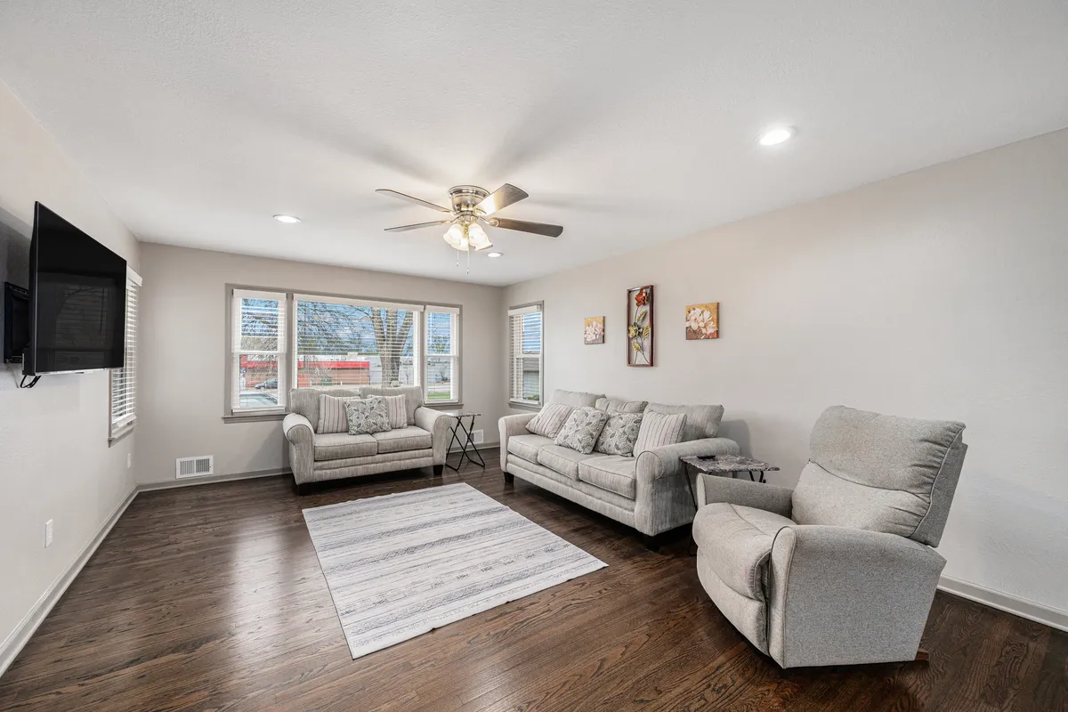 A comfortable sitting area with plush sofas and a recliner set around a modern rug and coffee table, with sunlight streaming through the windows. The setup includes a television for entertainment and is arranged to facilitate conversation or relaxation. The overall mood is cozy and inviting, ideal for morning coffee, reading, or gathering with friends and family.