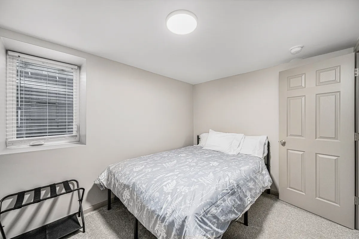 A tranquil bedroom featuring a bed with floral patterned bedding beside a small window with blinds. The setup includes a door leading to another room, creating a cozy, lived-in feel. Natural light gently filters in, making the corner perfect for a peaceful reading nook or quiet reflection.